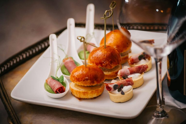 An elegant display of gourmet appetizers including mini burgers and prosciutto rolls served on a square plate.
